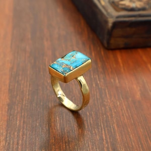 May include: A gold-colored ring featuring a rectangular turquoise stone with gold veining. The ring has a simple band and a rectangular setting that holds the stone. The ring is displayed on a wooden surface.