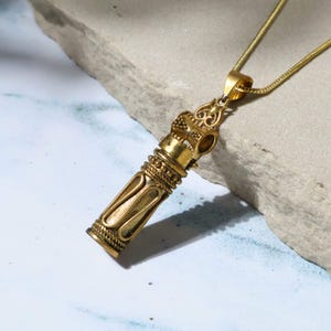May include: A gold-colored pendant necklace with a cylindrical design. The pendant has intricate detailing and is suspended from a delicate gold chain. The necklace is displayed on a light gray stone surface.