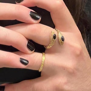May include: A gold-toned open ring shaped like a snake with textured scales and two black oval stones set in the head and tail, paired with a thin gold-toned band featuring a repeating diagonal etched pattern on the adjacent finger.