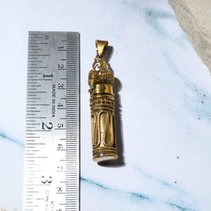May include: A small, ornate brass pendant with a cylindrical body and intricate detailing. The pendant is approximately 2.5 inches long, featuring a loop at the top for a chain. The design includes decorative patterns and a textured surface.