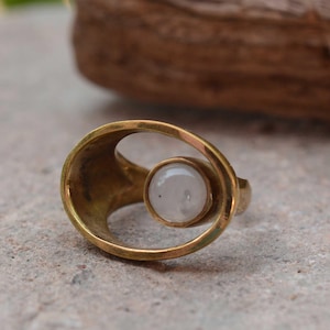 Könnte beinhalten: Ein goldfarbener Ring mit kreisförmigem Design, mit einem zentralen, runden, weißen Edelstein. Der Ring hat eine offene, fließende Form. Der Ring ist vor einem neutralen Hintergrund platziert.