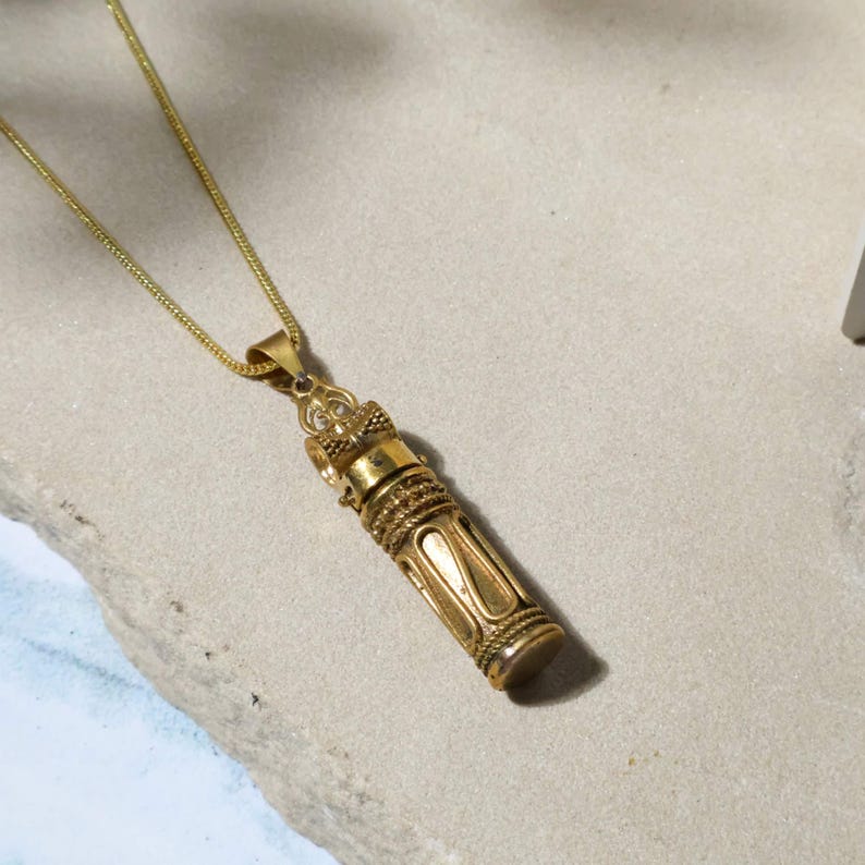 May include: Gold-colored pendant necklace featuring a cylindrical charm with intricate embossed designs. The charm has a decorative top and bottom, and is suspended from a delicate gold chain. The pendant is likely made of metal and has a vintage or antique aesthetic.