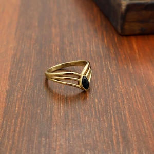 May include: A gold-colored ring with a black oval-shaped stone. The ring has a split band design with three parallel lines on each side of the stone. The ring is set on a wooden surface.