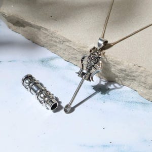 Könnte beinhalten: Eine silberfarbene Halskette mit Anhänger und einem detaillierten, verzierten Design. Der Anhänger ist an einer feinen Kette befestigt. Neben dem Anhänger befindet sich ein silberfarbener zylindrischer Gegenstand mit dekorativen Mustern.