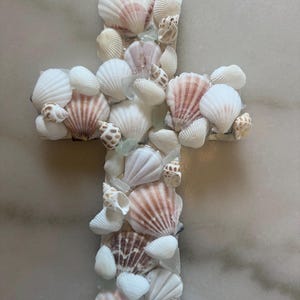 May include: A decorative cross crafted from various seashells, including white and pink scallop shells and small spiral shells. The shells are arranged on a white base, creating a textured surface. The cross is a coastal-themed decorative item.