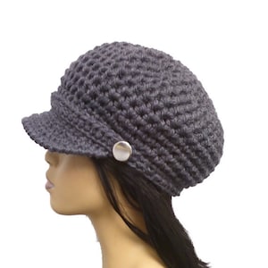 Puede incluir: Un gorro de punto gris con visera y un botón plateado. El gorro se lleva en un maniquí con el pelo largo y negro.