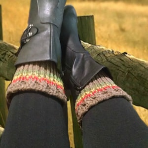May include: A pair of black leather boots with brown and beige knitted leg warmers. The boots are resting on a wooden fence.