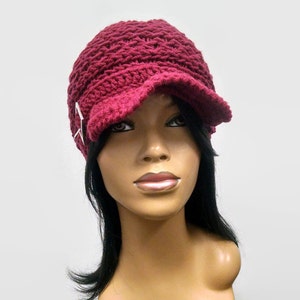 May include: A burgundy crocheted beanie with a visor and a silver buckle. The beanie is on a mannequin head with black hair.