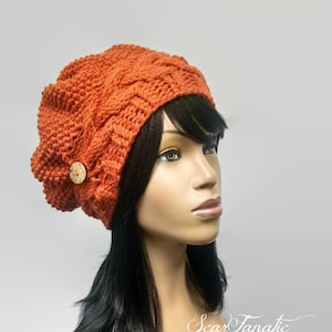 Puede incluir: Un gorro de punto naranja brillante con un patrón de punto de cable y un botón de madera. El gorro es holgado y tiene un ajuste relajado.