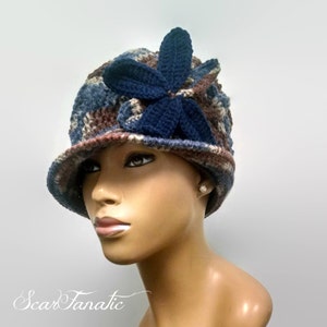 May include: A brown, blue, and beige crocheted hat with a blue crocheted flower on top. The hat is on a mannequin head.