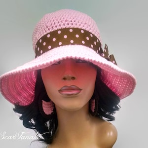 May include: A pink crochet hat with a wide brim and a brown ribbon with white polka dots. A matching bow is on the side. The hat is part of a set with matching earrings.