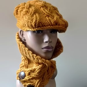 Puede incluir: Un gorro y un cuello a juego de punto amarillo mostaza con un patrón de punto trenzado y detalles de botones de madera.