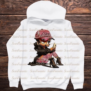 May include: White hoodie featuring a cartoon illustration of a character wearing a red Aries cap and patterned pants, holding a phone. The design includes the word "Aries" on the cap and is set against a wooden background.