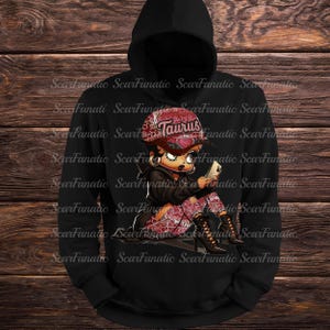May include: Black hoodie featuring a cartoon illustration of a woman wearing a red Taurus cap, black jacket, and patterned pants. The woman is holding a phone. The hoodie has the text "ScarFanatic" repeated.