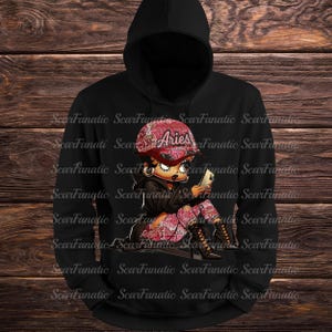 May include: Black hoodie featuring a cartoon illustration of a woman wearing a pink Aries hat and matching pants, holding a phone. The design includes a black jacket and high-heeled boots. The background is a wooden texture.
