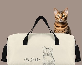 Peut inclure: Sac de sport crème avec des poignées noires et une bandoulière noire. Le sac présente un croquis de chat et le texte "My Bubba". Un chat tabby brun et noir est perché sur le sac.
