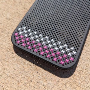 May include: A black rectangular phone case featuring a grid of small circular perforations. A cross-stitch pattern is embroidered through the holes, transitioning from white stitches at the top to vibrant pink stitches at the bottom corner.