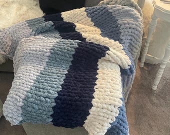 Puede incluir: Una manta de punto grueso en tonos azules, blancos y grises. La manta está sobre un sofá gris, mostrando su diseño texturizado y a rayas. El hilo grueso crea una estética acogedora e invitadora, ideal para la decoración del hogar.