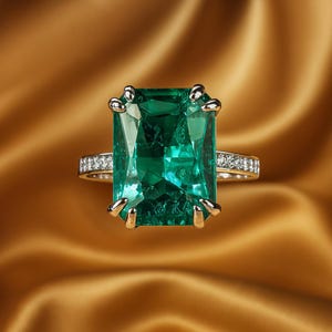 Könnte beinhalten: Ein goldener Ring mit einem großen, rechteckigen Smaragd-Edelstein. Der Smaragd hat eine leuchtend grüne Farbe und wird von goldenen Krappen gehalten. Das Ringband ist mit kleinen, funkelnden Diamanten besetzt.