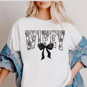 May include: White t-shirt with the word "WIFFEY" in a floral design, with a black bow at the bottom. The shirt is made of a soft material and is suitable for casual wear.
