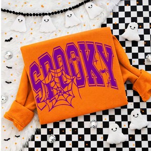 Może przedstawiać: Pomarańczowa bluza z napisem SPOOKY wydrukowanym dużymi fioletowymi literami w stylu uniwersyteckim. Projekt zawiera grafikę pajęczyny wkomponowaną w litery oraz małe ilustracje pająków i gwiazdek wykonane fioletowym tuszem.