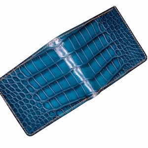 Könnte beinhalten: Rechteckige, türkisblaue Geldbörse mit Krokodilleder-Textur. Die Geldbörse hat ein zweifaches Design mit sichtbarer Naht und Nähten in einer kontrastierenden, hellbraunen Farbe. Der Hintergrund ist weiß.
