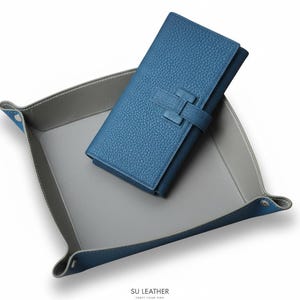 Puede incluir: Una cartera de cuero azul con superficie texturizada y cierre a juego descansa dentro de una bandeja de cuero bicolor gris y azul. La bandeja tiene esquinas redondeadas y está sobre un fondo blanco.
