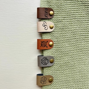 Könnte beinhalten: Nahaufnahme von fünf Lederetiketten in verschiedenen Farben, darunter Braun, Beige, Orange, Grau und Beige, die an einem hellgrünen Strickstoff befestigt sind. Jedes Etikett hat einen Messingverschluss und ein eingraviertes Design oder Text.