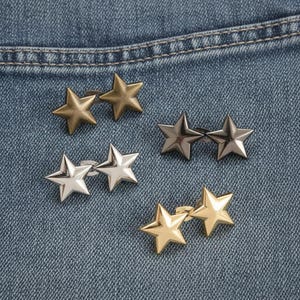 May include: Close-up of several pairs of star-shaped metal studs in various finishes: gold, silver, and bronze. The studs are arranged on a blue denim fabric, showcasing their different metallic tones and textures.