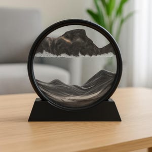 Peut inclure: Une pièce d'art de sable noire et circulaire avec un paysage de montagne. Le sable, dans des tons de gris et de noir, crée un effet visuel dynamique dans le verre. Posée sur un socle rectangulaire noir, pour un bureau ou une étagère.