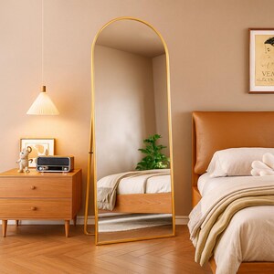 Peut inclure: Un miroir sur pied, de forme arquée et au cadre doré, est placé dans une chambre. Il reflète l'intérieur de la pièce. Une table de chevet en bois avec une lampe et une radio sont visibles. Le miroir mesure environ 170 cm de haut.