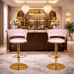 Pode incluir: Duas banquetas de bar de veludo rosa com estruturas e bases de metal dourado. As banquetas têm encosto tufado e assento redondo. O fundo apresenta um bar com garrafas de licor, copos e uma bancada de mármore.