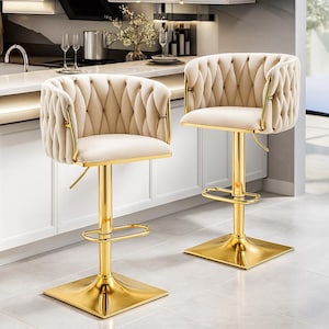Puede incluir: Dos taburetes de bar de terciopelo beige con marcos y bases de metal dorado. Los asientos tienen un diseño de respaldo acolchado y un asiento cuadrado. Los taburetes tienen un reposapiés y una palanca de ajuste de altura. Están en una cocina.