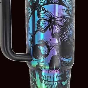 May include: Black travel mug with a skull and butterfly design. The skull is overlaid with iridescent blue, purple, and green butterflies. The mug has a black handle and a black lid with a metallic iridescent band.