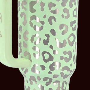 Puede incluir: Un vaso de acero inoxidable verde menta con asa, con un estampado de leopardo plateado. El vaso tiene una tapa transparente y un asa verde menta. El logotipo de la marca es visible en la parte inferior.