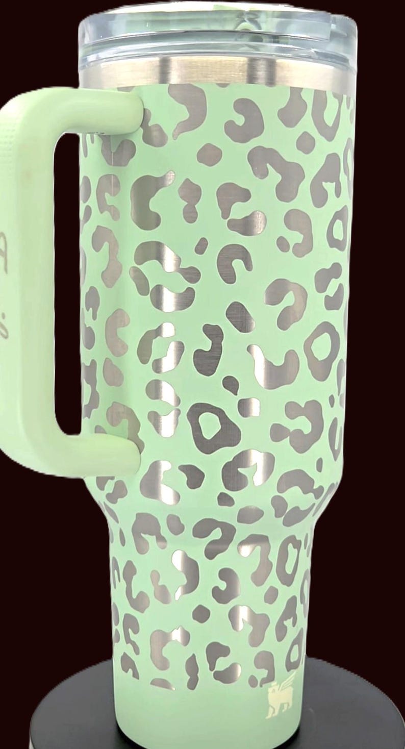 Puede incluir: Un vaso de acero inoxidable verde menta con asa, con un estampado de leopardo plateado. El vaso tiene una tapa transparente y un asa verde menta. El logotipo de la marca es visible en la parte inferior.