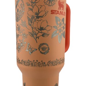 May include: A large, orange Stanley tumbler with a white lid and orange handle. The tumbler is decorated with silver floral and insect designs. The Stanley logo is visible on the side. Includes a straw.