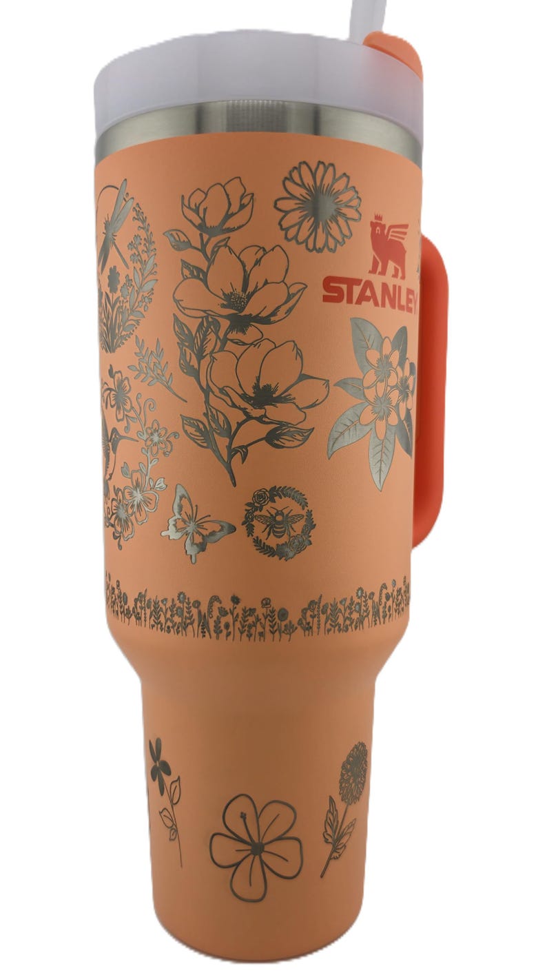May include: A large, orange Stanley tumbler with a white lid and orange handle. The tumbler is decorated with silver floral and insect designs. The Stanley logo is visible on the side. Includes a straw.