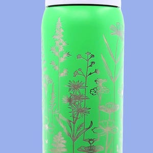 Pu&ograve; includere: Una borraccia Owala verde con un design floreale argentato. La borraccia ha una fascia bianca, un manico viola e un coperchio azzurro. La parola "owala" &egrave; stampata in bianco. La borraccia &egrave; cilindrica e sembra essere in metallo.