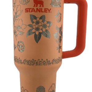 May include: A coral-colored Stanley tumbler with a floral design and the Stanley logo. The tumbler has a silver rim and a coral handle. The floral design includes various flowers and leaves in a silver color. The lid and handle are coral.