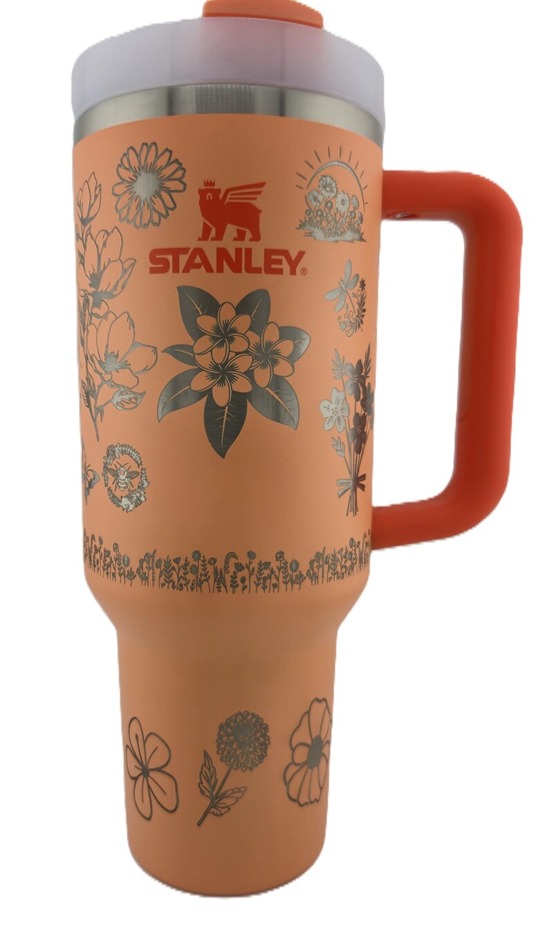 May include: A coral-colored Stanley tumbler with a floral design and the Stanley logo. The tumbler has a silver rim and a coral handle. The floral design includes various flowers and leaves in a silver color. The lid and handle are coral.