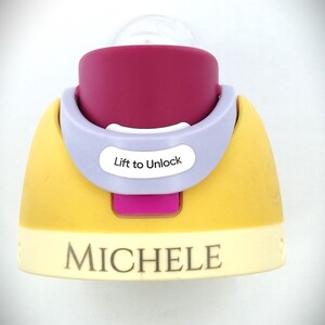 Pu&ograve; includere: Un biberon per bambini con tettarella trasparente e base viola, gialla e rosa. La base ha il nome "MICHELE" stampato sopra. Il biberon ha un colletto lilla chiaro con le parole "Lift to Unlock".