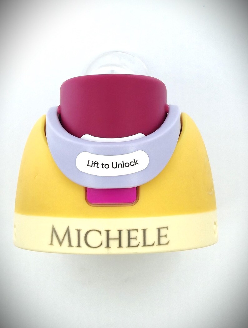 Pu&ograve; includere: Un biberon per bambini con tettarella trasparente e base viola, gialla e rosa. La base ha il nome "MICHELE" stampato sopra. Il biberon ha un colletto lilla chiaro con le parole "Lift to Unlock".