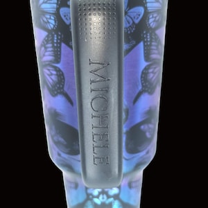Puede incluir: Vaso negro con asa, con un dise&ntilde;o de calavera con detalles de mariposas en tonos azules y morados. El nombre "MICHELE" est&aacute; grabado en el asa. El borde superior tiene un acabado met&aacute;lico azul.