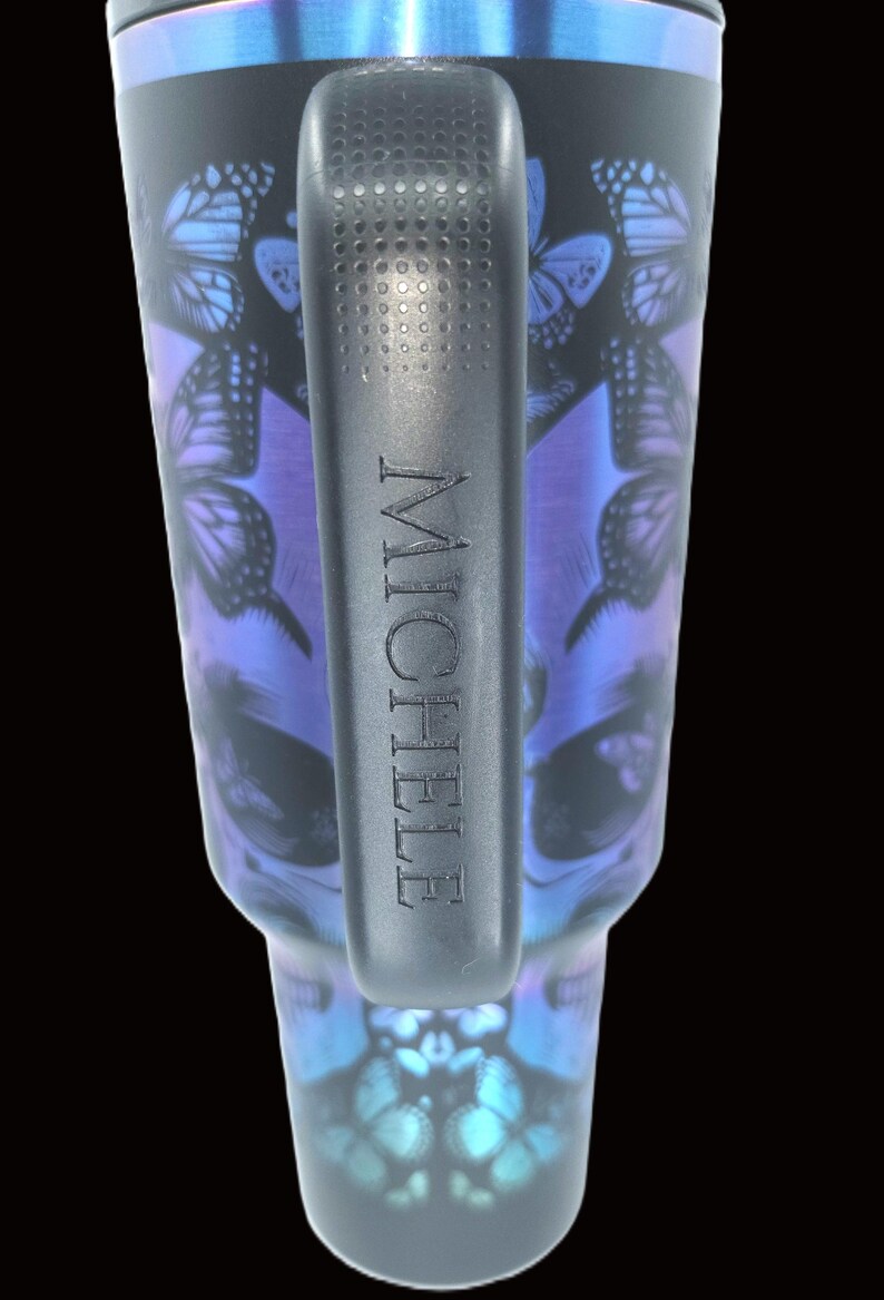 Puede incluir: Vaso negro con asa, con un dise&ntilde;o de calavera con detalles de mariposas en tonos azules y morados. El nombre "MICHELE" est&aacute; grabado en el asa. El borde superior tiene un acabado met&aacute;lico azul.