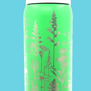 Pu&ograve; includere: Una borraccia verde con un design floreale. La bottiglia ha una parte superiore bianca, un coperchio viola chiaro e il marchio "owala" stampato sul lato. Il design floreale &egrave; argentato e presenta varie piante.