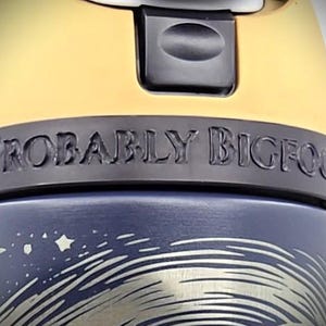 Pu&ograve; includere: Primo piano di una tazza da viaggio blu e gialla. La tazza presenta la scritta "PROBABLY BIGFOOT" in una fascia grigio scuro. La parte inferiore della tazza ha una grafica argentata di un cielo notturno con stelle e un design a spirale.