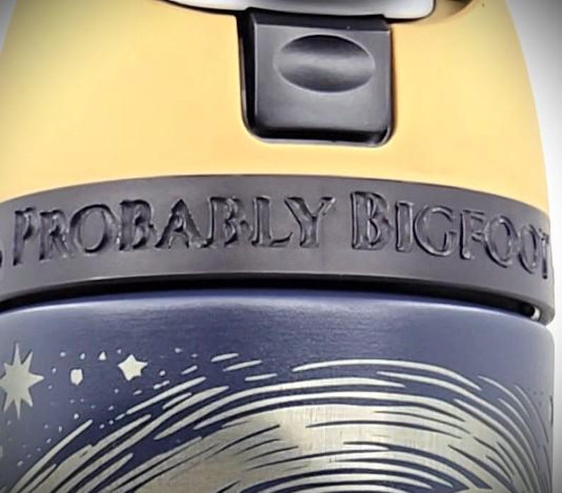 Pu&ograve; includere: Primo piano di una tazza da viaggio blu e gialla. La tazza presenta la scritta "PROBABLY BIGFOOT" in una fascia grigio scuro. La parte inferiore della tazza ha una grafica argentata di un cielo notturno con stelle e un design a spirale.