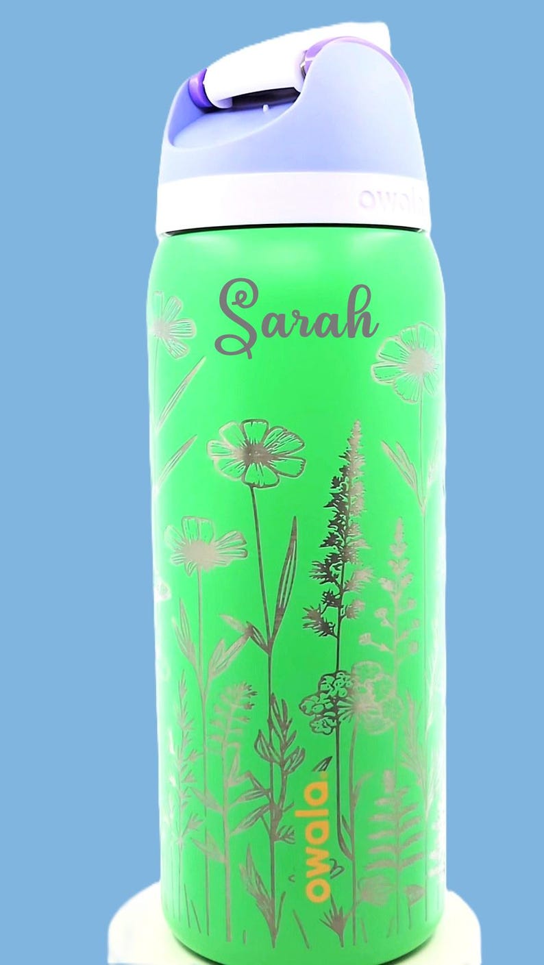 Pu&ograve; includere: Borraccia verde con un design floreale e il nome "Sarah" in corsivo. La bottiglia ha una parte superiore bianca e un manico viola. Il marchio "owala" &egrave; stampato sulla bottiglia.