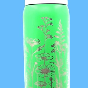 Pu&ograve; includere: Una borraccia verde con un design floreale argentato. Il tappo &egrave; viola e riporta la scritta "Lift to Unlock". Il design floreale presenta vari fiori e piante.